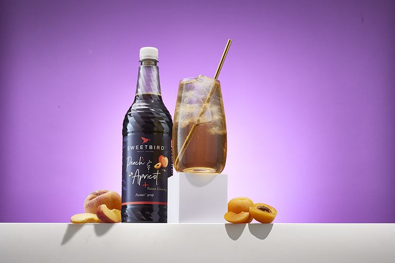 Sweetbird Peach & Apricot syrup bottle next to a glass of iced peach-colored drink with a straw, surrounded by peaches and apricots on a white surface.