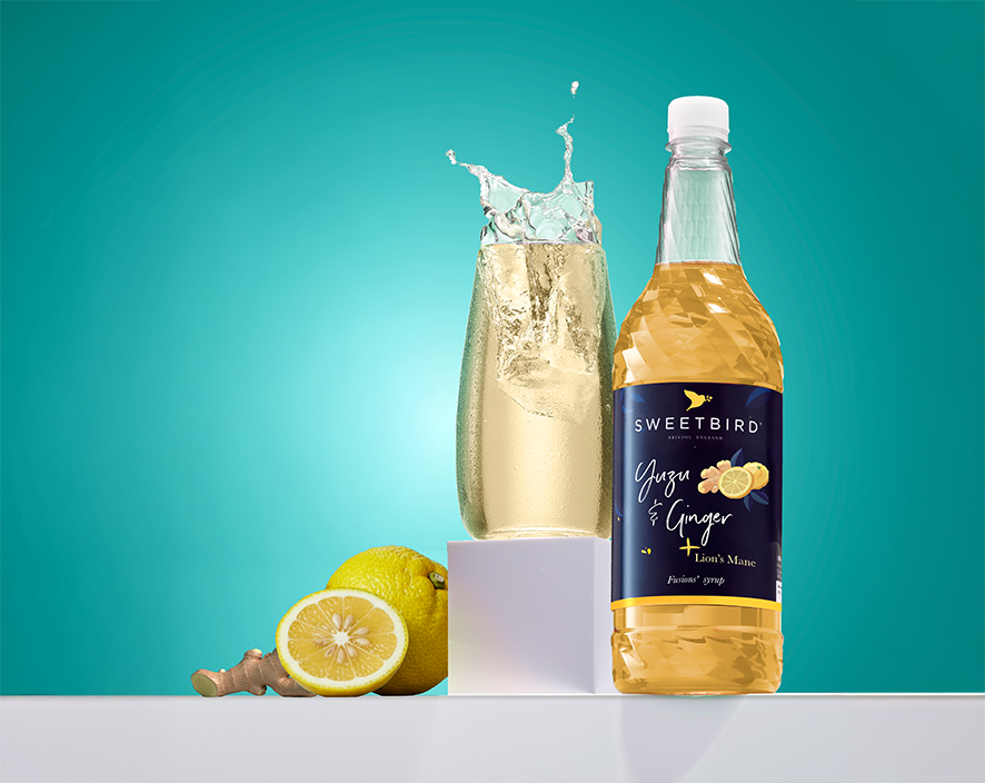 Bottle of Sweetbird Yuzu & Ginger fusion syrup next to a tall glass of iced drink splashing at the rim, with fresh yuzu citrus and ginger on a white surface.